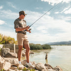 Man fishing by a lake with mountains in the background while wearing Mossy Oak and Ridge Belt gear.